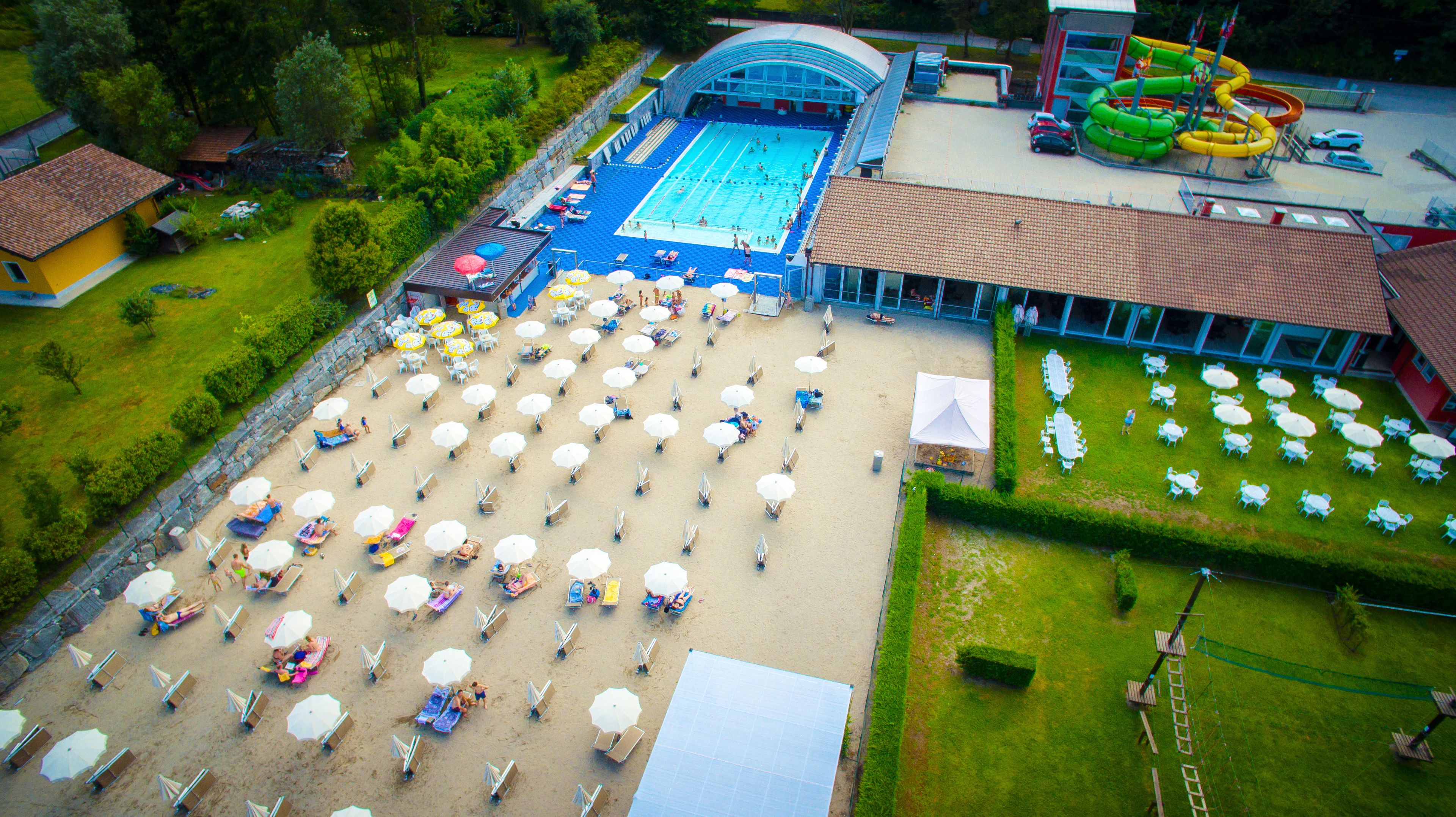 An aerial view of a swimming pool area with multiple sun loungers and umbrellas, a covered seating area, and a water slide.