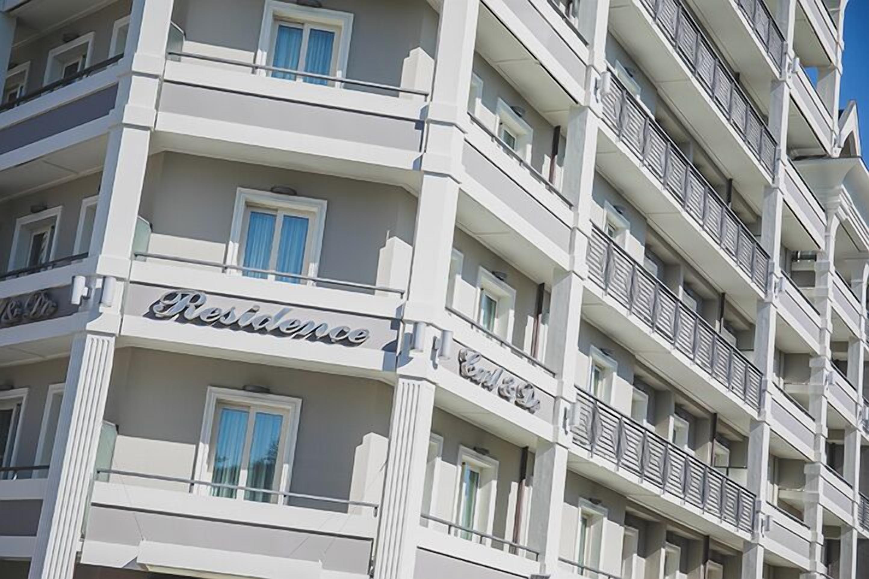 Un hôtel à plusieurs étages doté de balcons, une enseigne indiquant « Résidence », et un ciel bleu clair en arrière-plan.