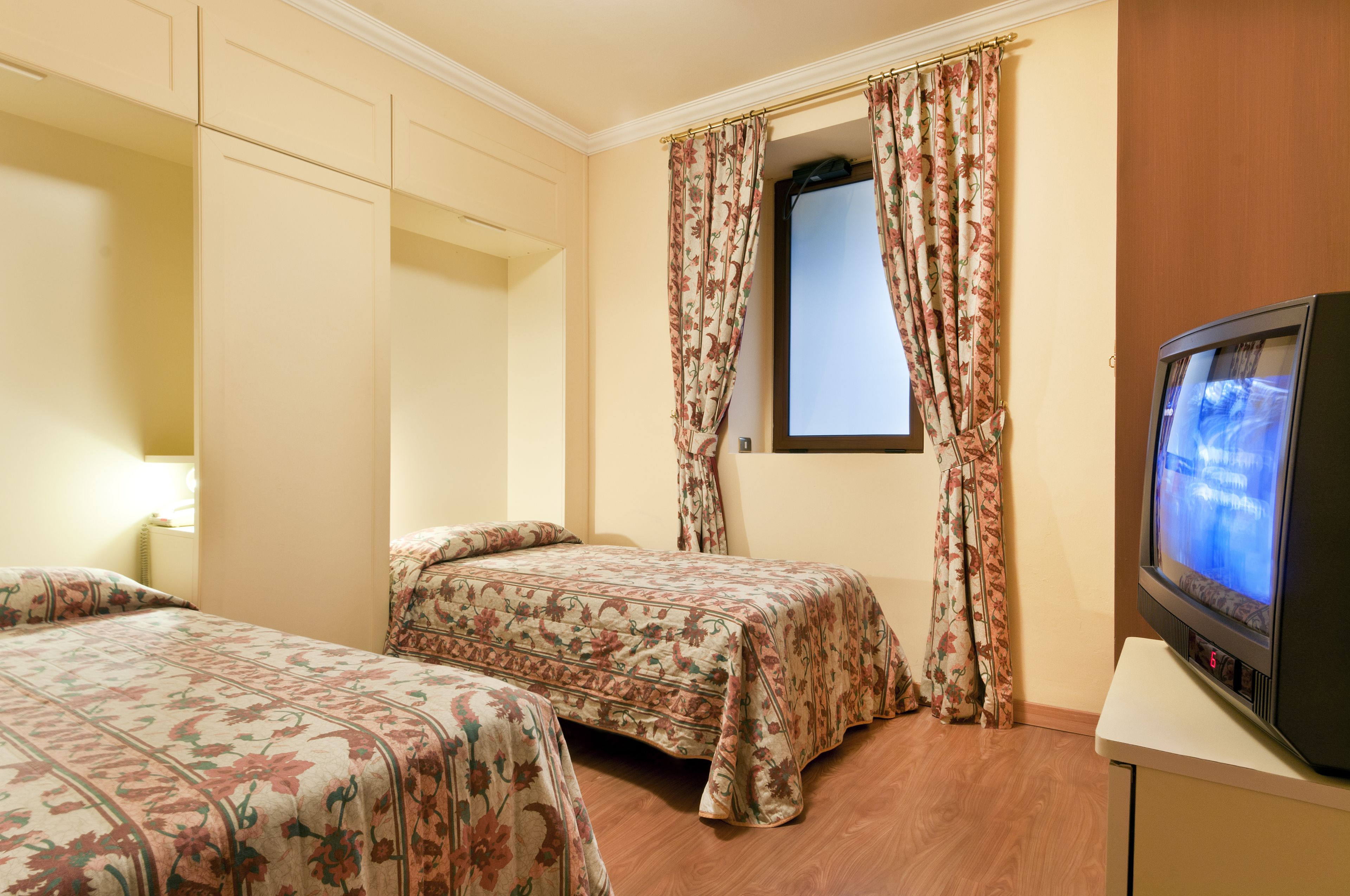 A hotel room with two beds, a television, and a window with floral curtains.