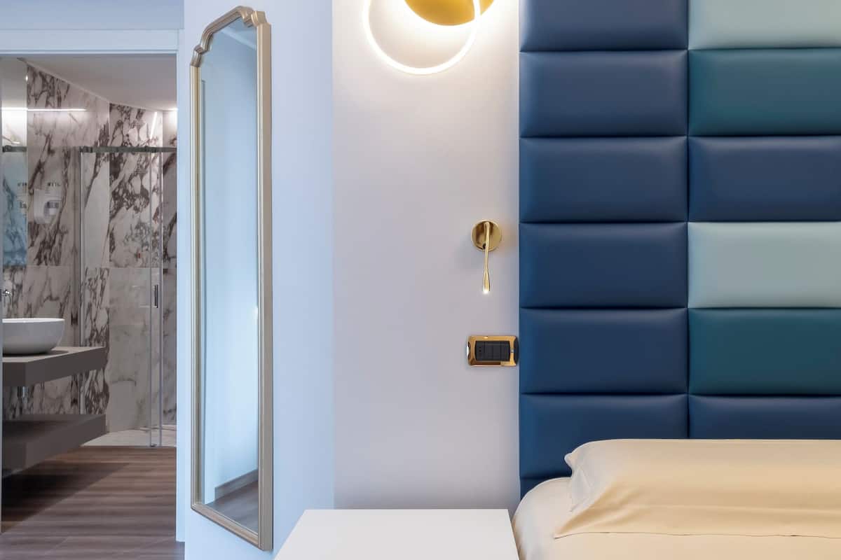 Un dormitorio moderno con cabecera azul, una mesita de noche con cajón, un espejo grande y una lámpara de pared.