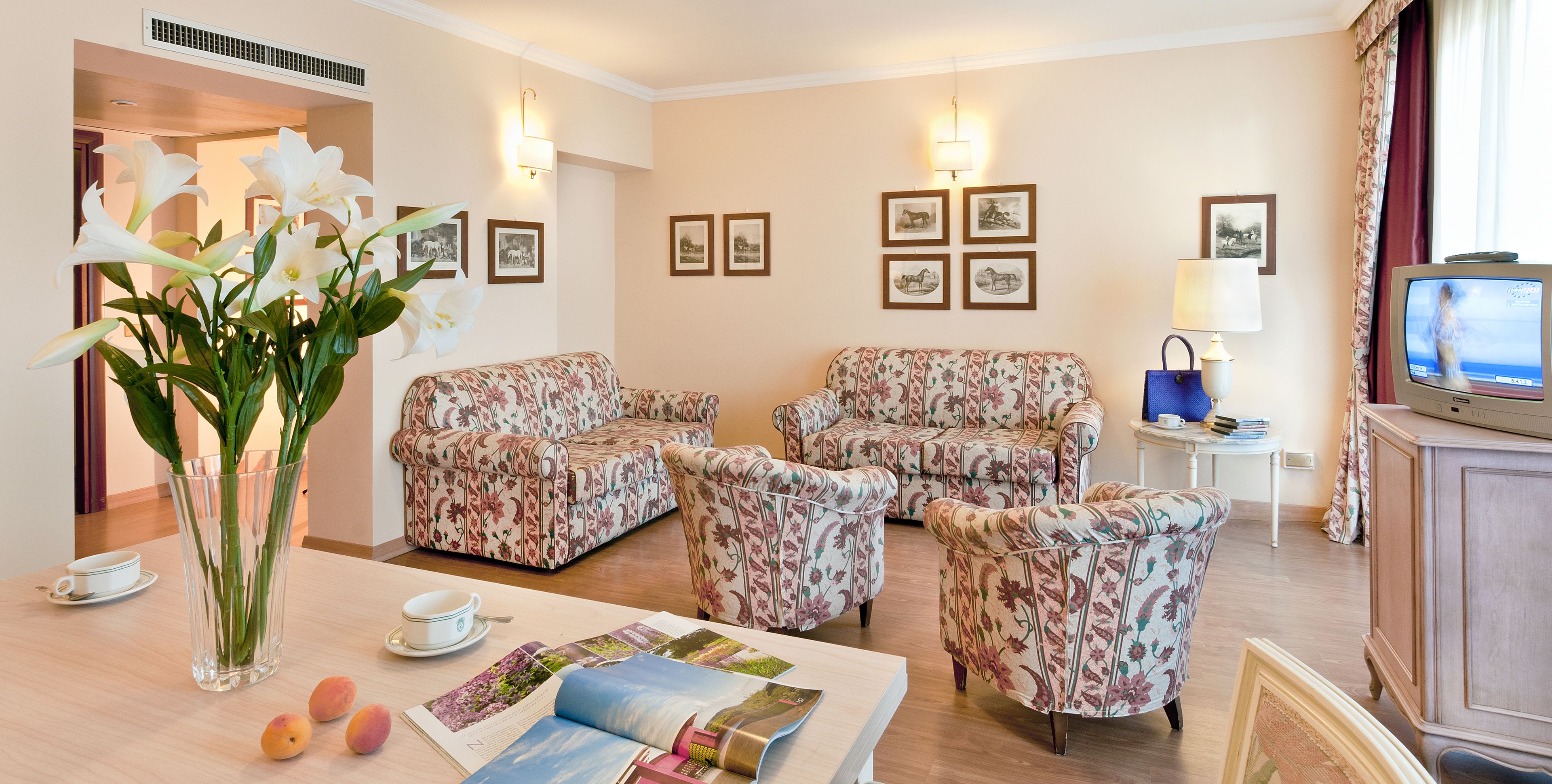 A living room with floral-patterned sofas, a coffee table with magazines, a vase with flowers, and framed pictures on the wall.