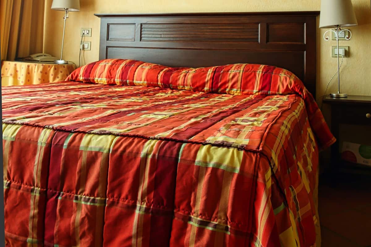 A hotel room with a wooden ceiling, a bed with a red and yellow plaid bedspread, two bedside lamps, and a nightstand with a lamp.