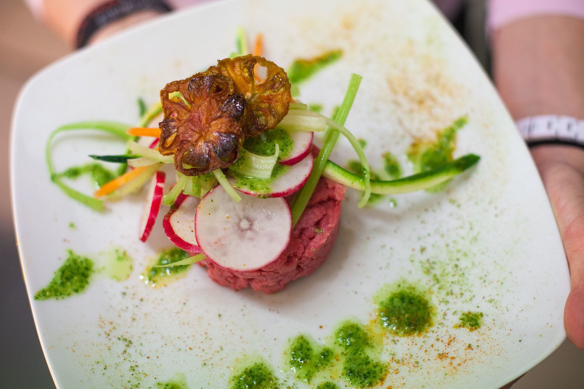 A square plate with a meat-based dish, garnished with vegetables and a green sauce.