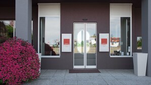 L'ingresso di un hotel con una porta in vetro, incorniciato da due finestre, e un piccolo giardino con fiori rosa sulla sinistra.