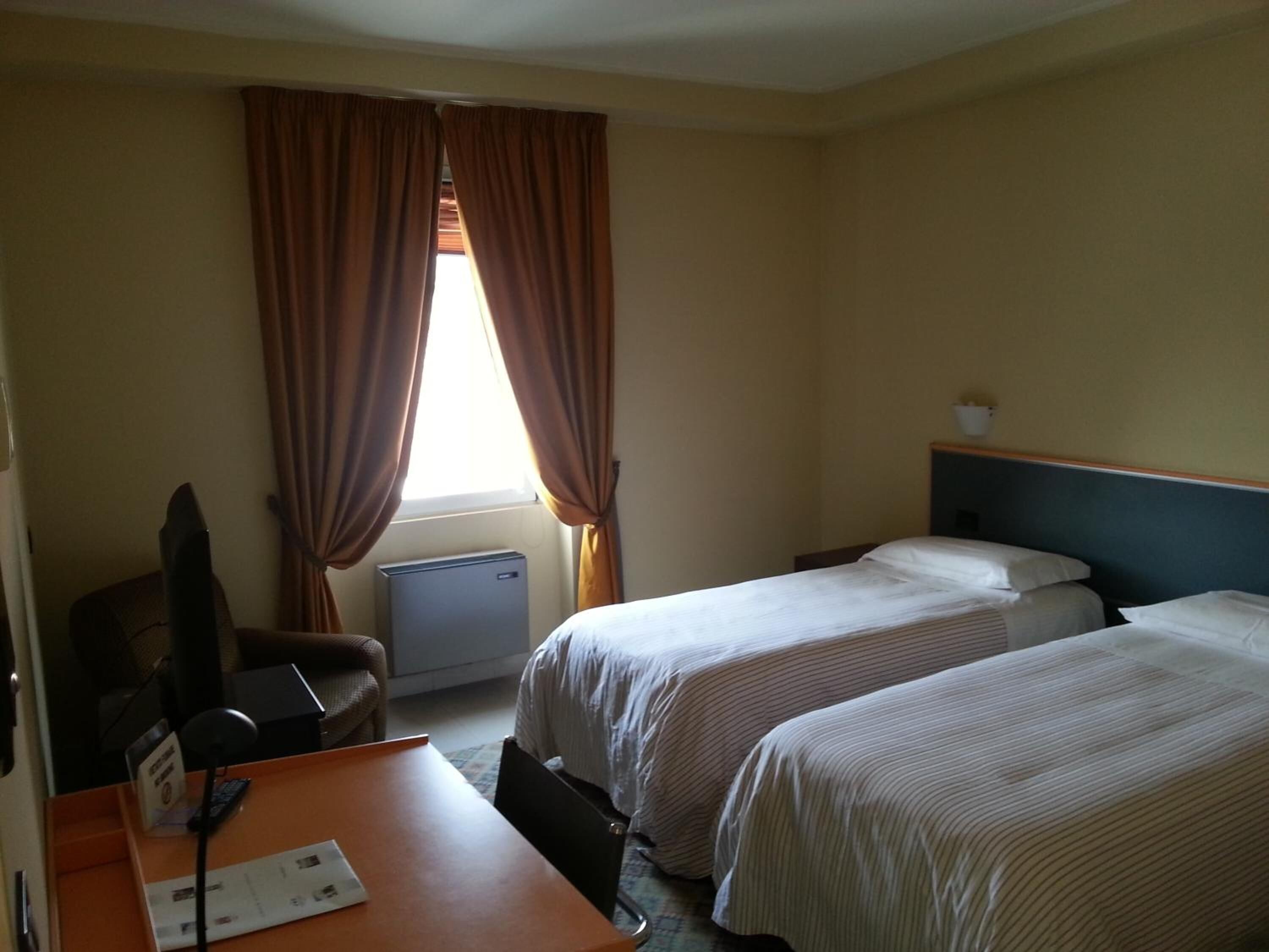 A hotel room with two beds, a desk with a chair, a small table, and a window with curtains.