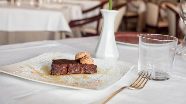 Un piattino con torta al cioccolato e noci, un vaso bianco con un fiore giallo e un bicchiere di vetro trasparente su una tovaglia bianca.