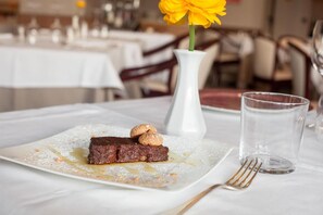 A dessert plate with a chocolate cake and nuts, a white vase with a yellow flower, and a clear glass on a white tablecloth. - Hotel La Bettola (Carisio)