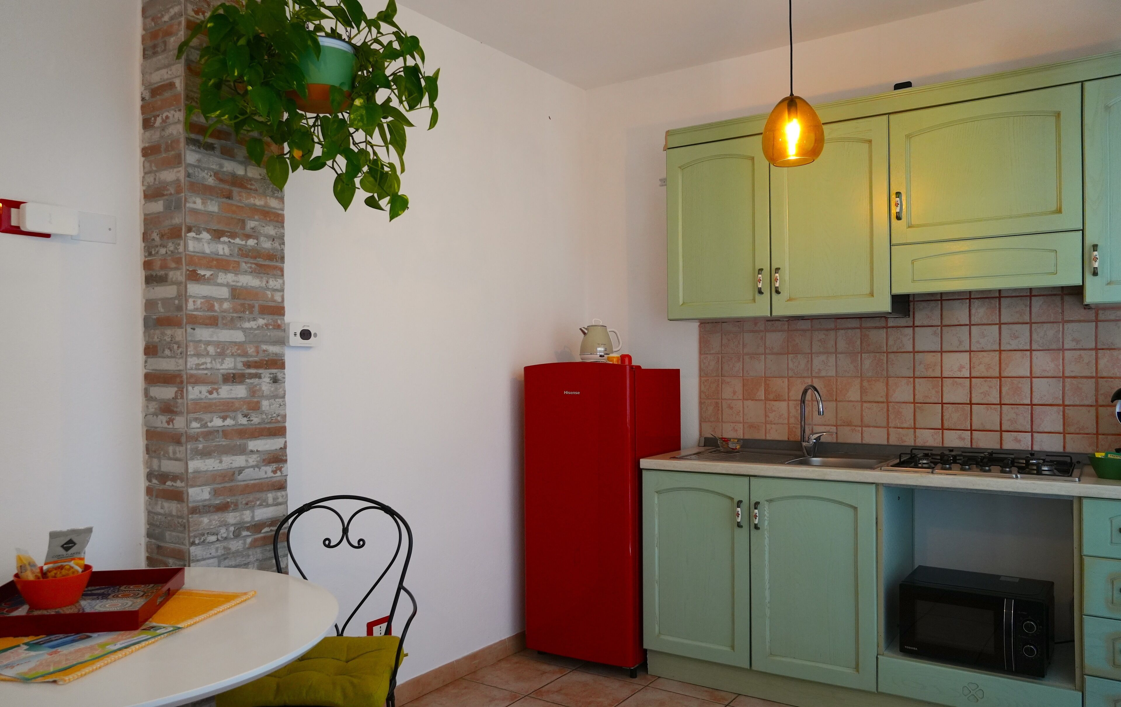 Una cocina con armarios verdes, un refrigerador rojo, una planta colgante, una pared blanca y un salpicadero de azulejos.