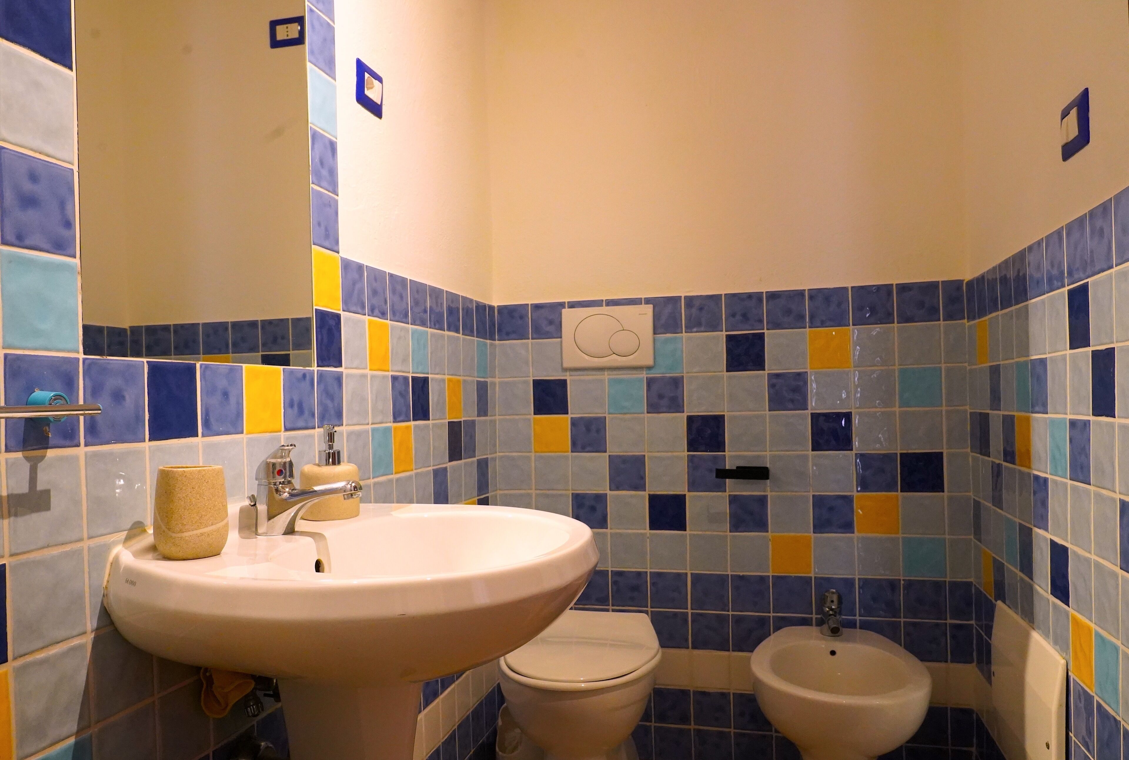 Une salle de bain avec un lavabo, des toilettes et un bidet, dont le carrelage est bleu, jaune et blanc.