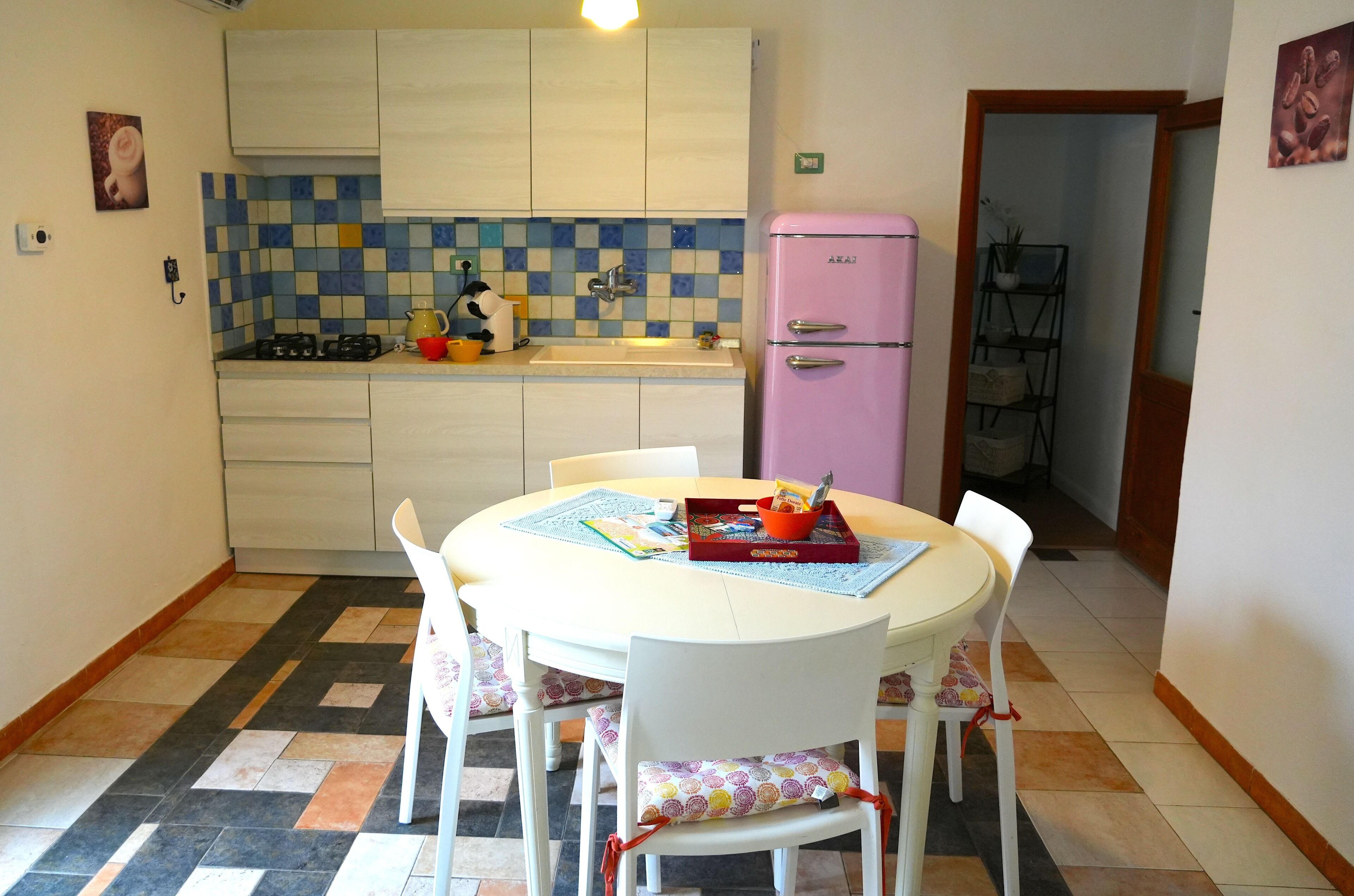 Een kleine keuken met een betegelde spatwand, een roze koelkast, en een eethoek met een tafel en stoelen.