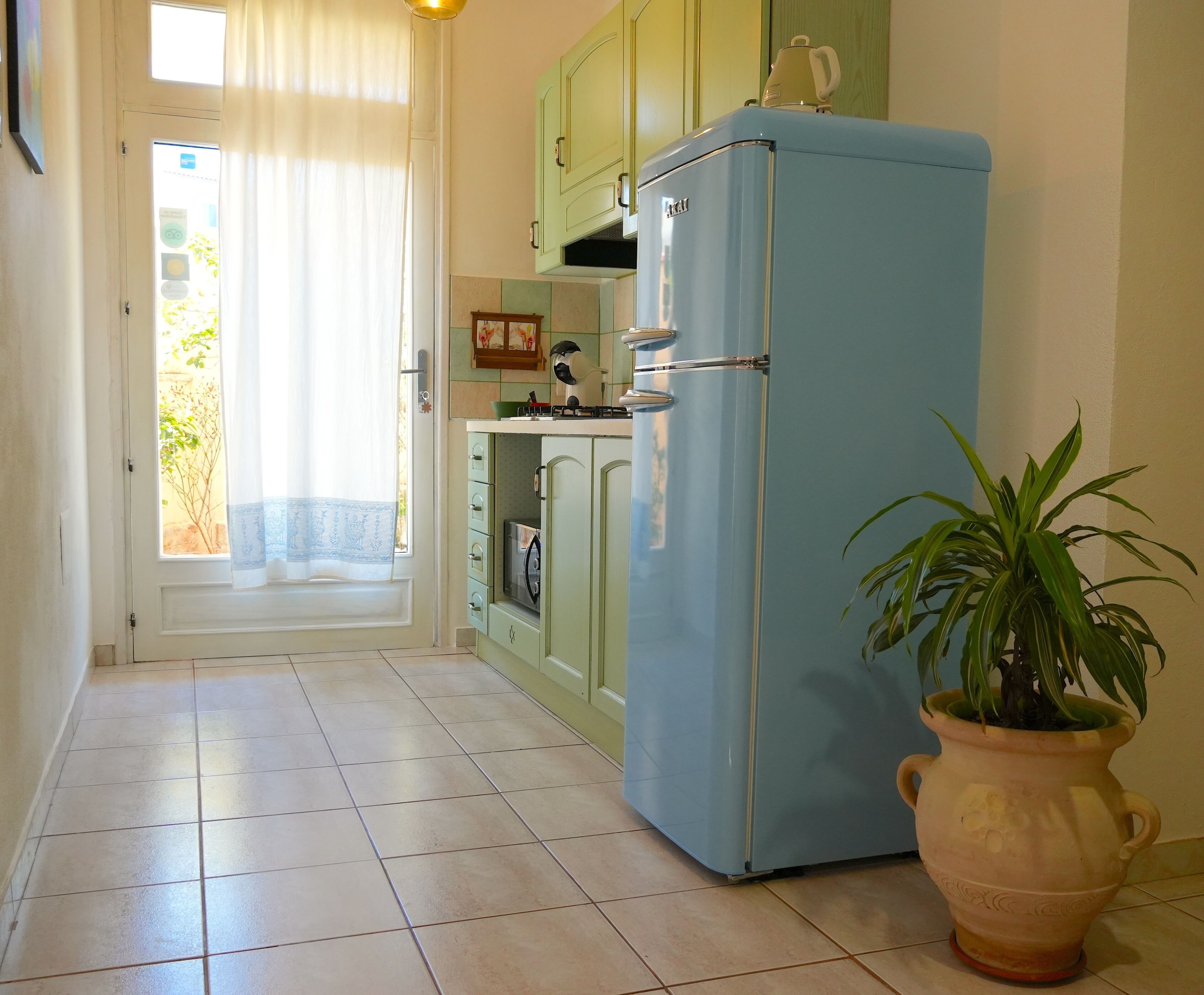 A kitchen with a blue refrigerator, green cabinets, a tiled floor, and a potted plant.