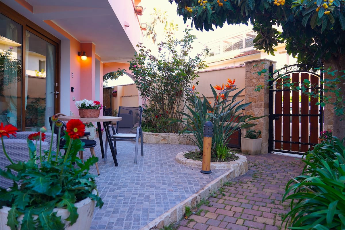 Un patio al aire libre con una mesa y sillas, rodeado de plantas en macetas y un muro de piedra.