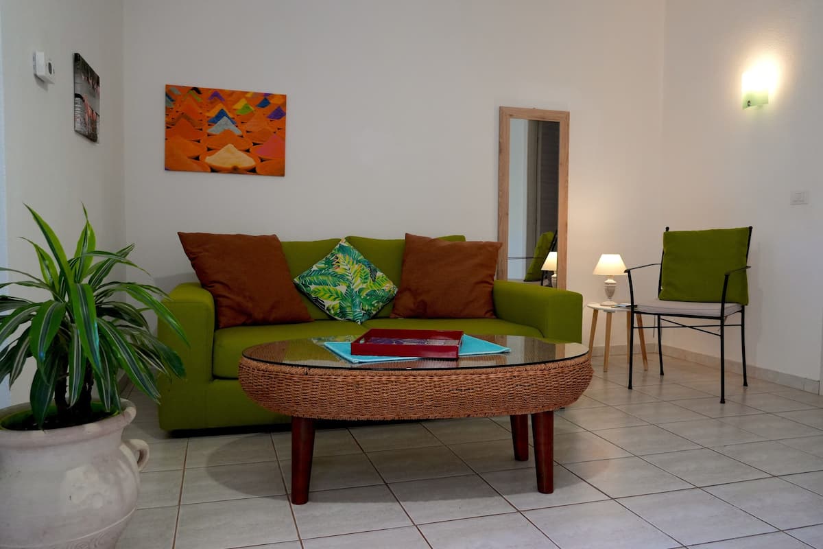 Ein Wohnzimmer mit einem grünen Sofa, einem Korbtisch, einem grünen Sessel, einer Topfpflanze und einem farbenfrohen abstrakten Gemälde.