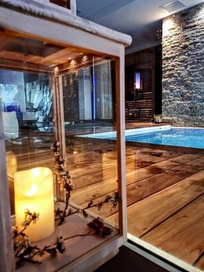 Terraza de madera con cerramiento de vidrio que contiene una vela y flores secas, con vistas a una piscina y un muro de piedra.