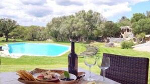 A table set for a meal with a bottle of wine, glasses, and plates of food, overlooking a swimming pool and a natural landscape. - Tenuta Pilastru (Arzachena)