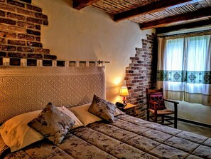 A bedroom with a brick wall, a bed with floral bedding, a nightstand with a lamp, and a chair by a window with curtains. - Tenuta Pilastru (Arzachena)