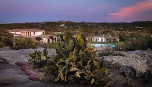 Paisaje desértico con un cactus grande al frente, una piscina y un edificio con múltiples arcos al fondo.