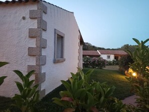 A white building with a stone facade and a window. There are plants in the foreground and a lit area in the background. - Tenuta Pilastru (Arzachena)
