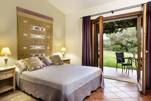 A bedroom with a large bed, two bedside tables with lamps, a patterned wall hanging, and a sliding glass door leading to an outdoor area with a table and chairs. - Tenuta Pilastru (Arzachena)