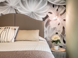 Un dormitorio con una cama, almohadas, una mesita de noche con un florero y una pared con un mural floral.