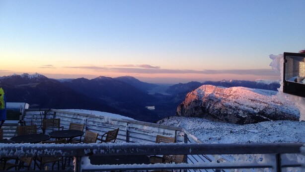 Zona de estar al aire libre cubierta de nieve con mesas y sillas, con vistas a un paisaje montañoso durante el atardecer.