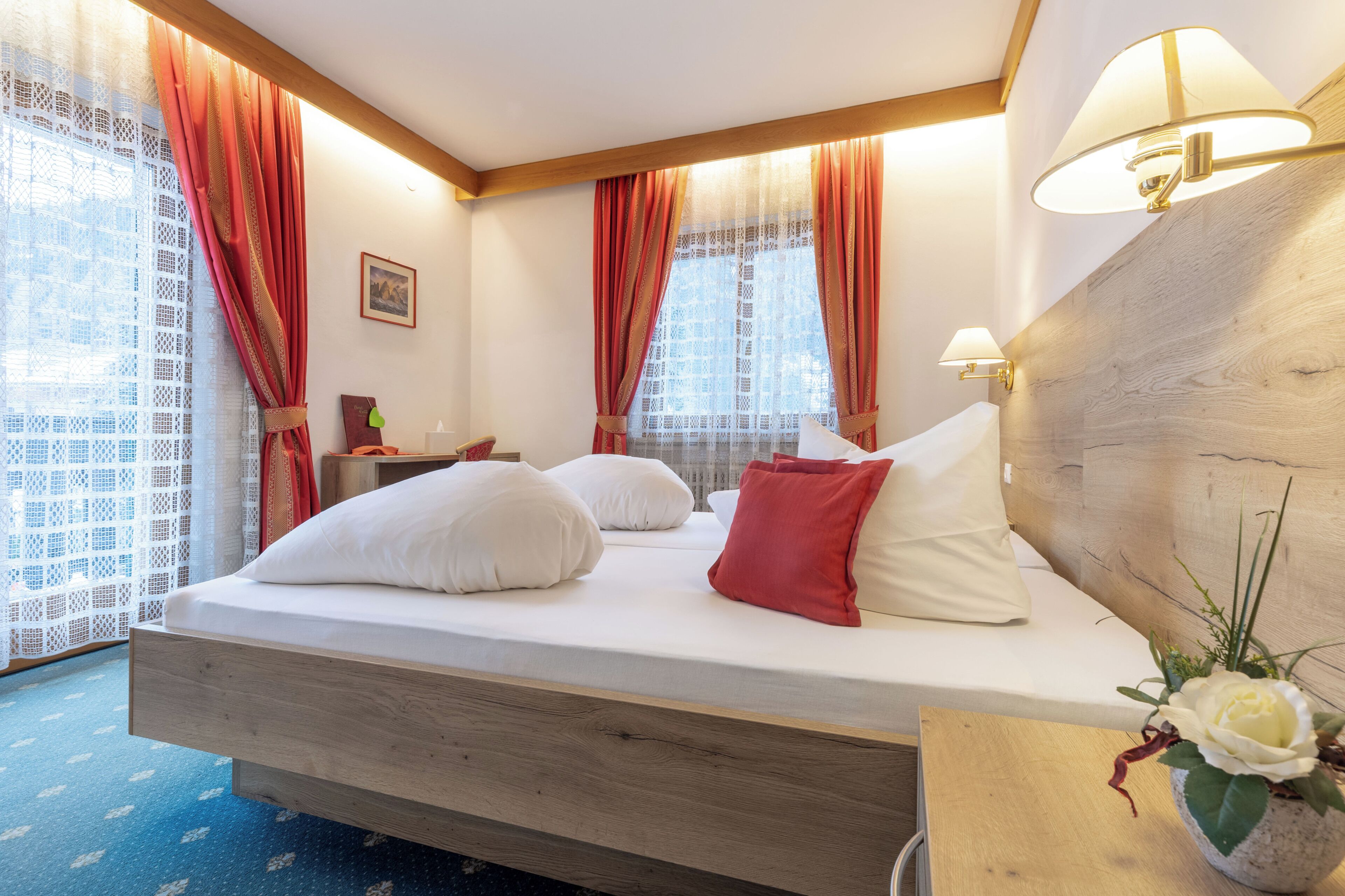 A hotel room with a wooden bed, red curtains, a bedside table with a lamp, and a window with a view.