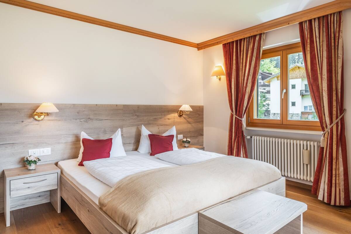 Een slaapkamer met een houten hoofdbord, twee nachtkastjes met lampen, een bed met wit en rode beddengoed, en een raam met gordijnen.