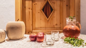 A wooden cabinet with a decorative diamond-shaped mirror, a wooden cutting board, two apples, a pitcher with mint leaves, and three glasses on a patterned tablecloth.
