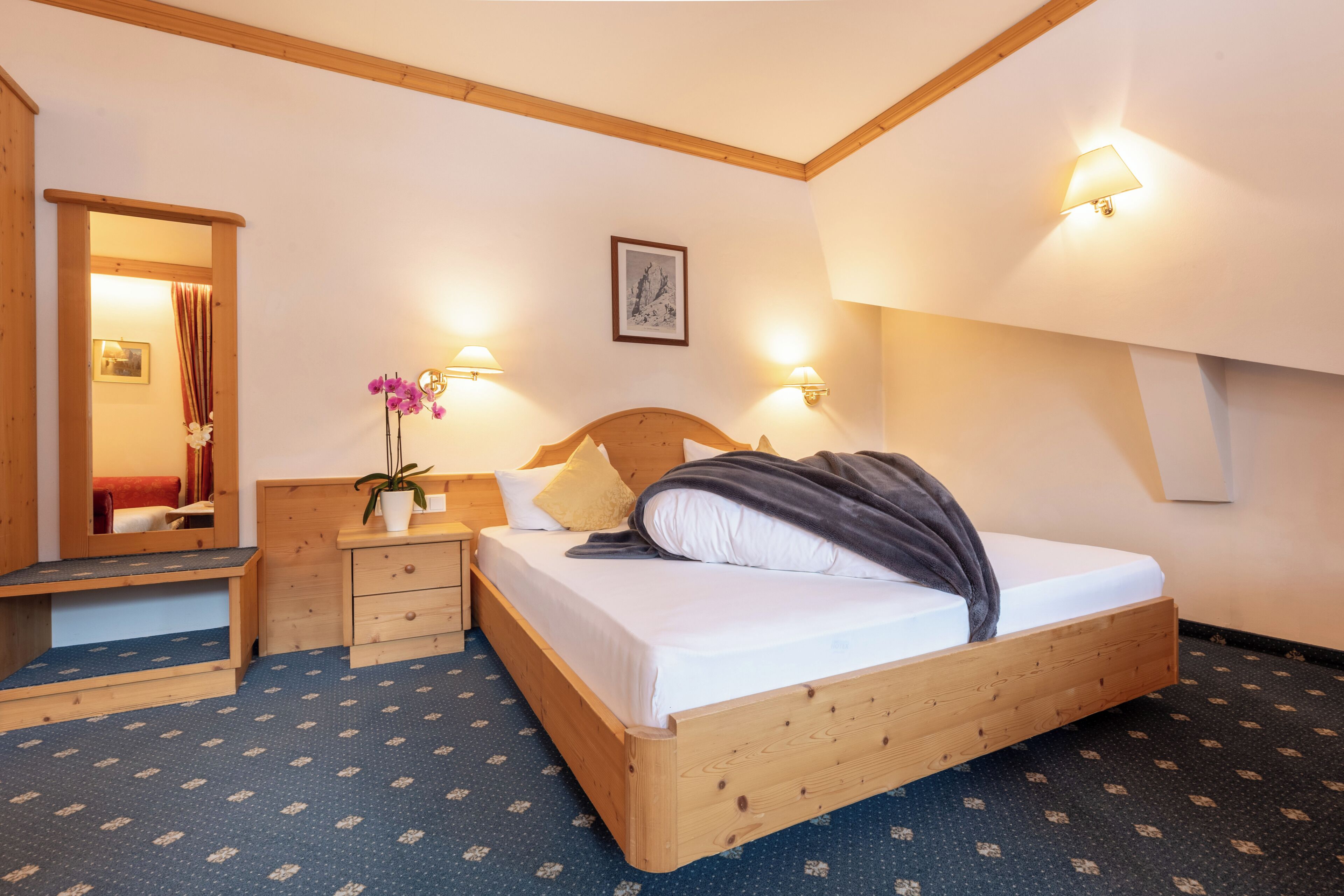 Ein Schlafzimmer mit einem hölzernen Bett, Nachttischen, einem Spiegel, Wandlampen und einem gerahmten Bild an der Wand.