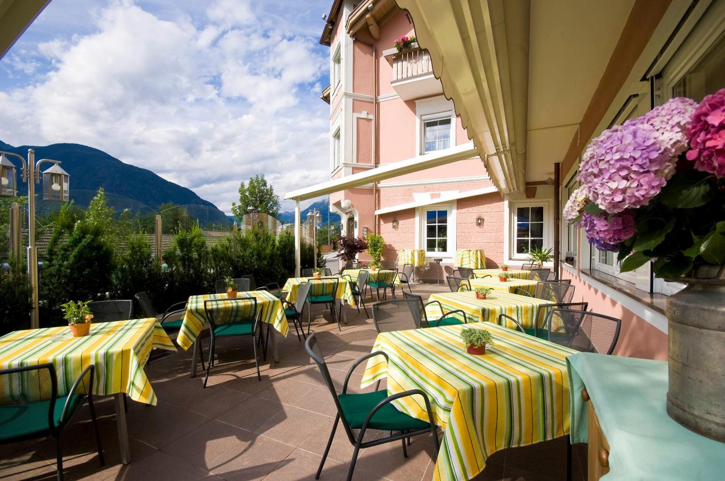 Une terrasse extérieure avec des tables et des chaises, un bâtiment à la façade rose et une montagne en arrière-plan.