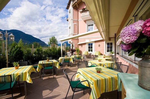 An outdoor patio with tables and chairs, a building with a pink facade, and a mountain in the background.