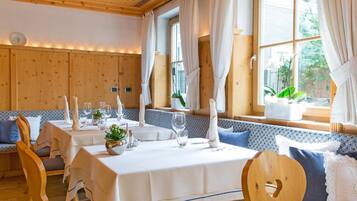 Zona pranzo con tavoli e panche in legno, tovaglie bianche e luce naturale proveniente da ampie finestre.