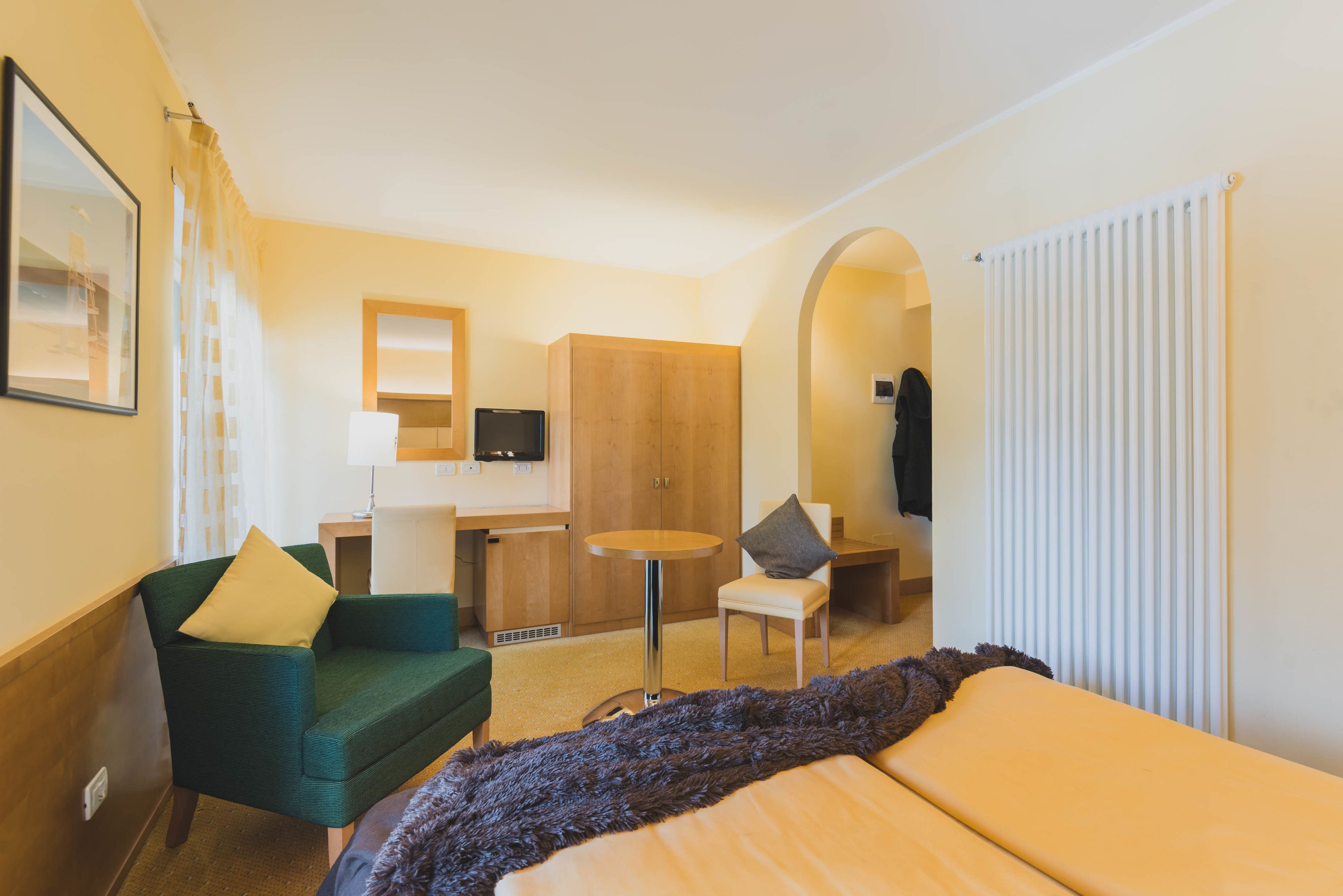 A hotel room with a bed, a green armchair, a small round table, a wooden wardrobe, a desk with a chair, and a television.