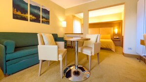 Quarto de hotel com um sofá verde, duas poltronas, uma mesa redonda e uma cama com cabeceira.