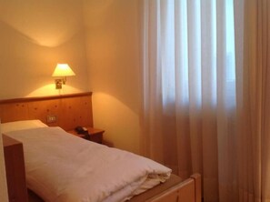 Una habitación de hotel con cama, mesita de noche, lámpara y ventana con cortinas.