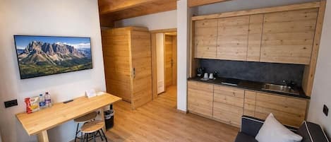 A compact living space with a wooden dining table, a sofa, a kitchen area, and a mounted TV displaying a mountain landscape.