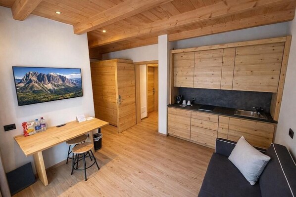 Un espacio habitable compacto con una mesa de comedor de madera, un sofá, una zona de cocina y un televisor montado que muestra un paisaje de montaña.