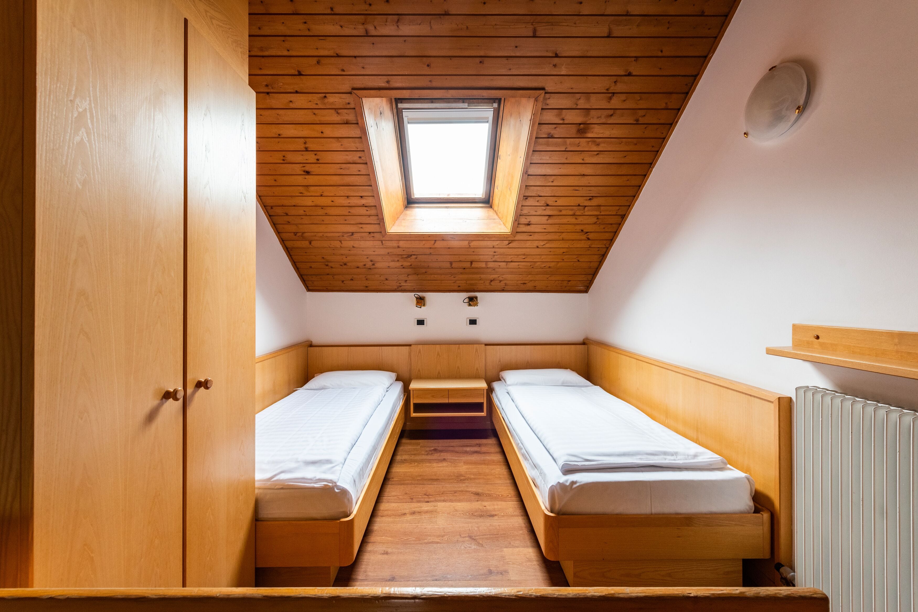 A small, compact room with two wooden beds, a skylight, and a wooden wardrobe.