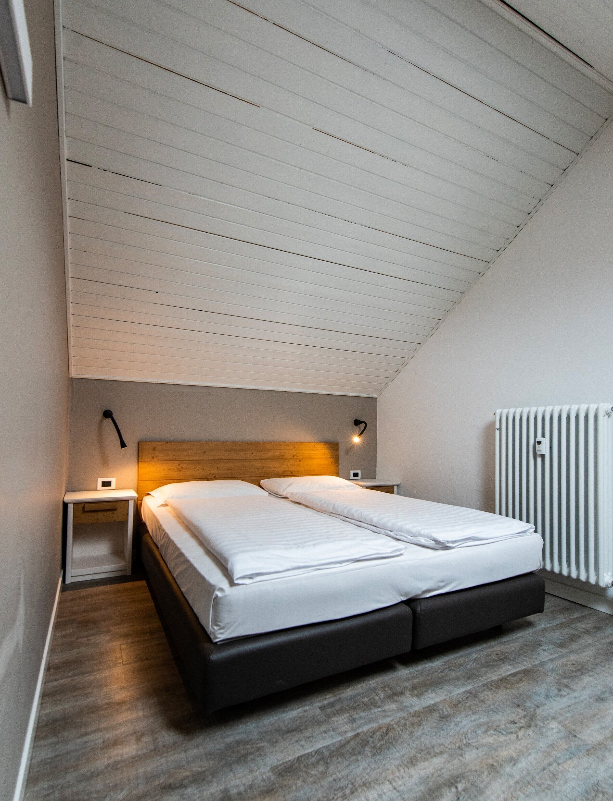 Ein Schlafzimmer mit schrägen Wänden, einem Bett mit weißer Bettwäsche, zwei Nachttischen mit Lampen und einem Heizkörper.