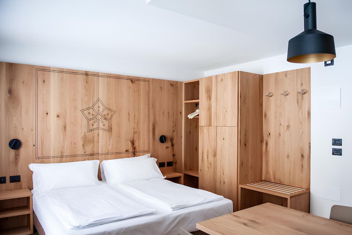 A modern hotel room with a wooden headboard, a bed with white linens, a wooden desk, and a pendant light.