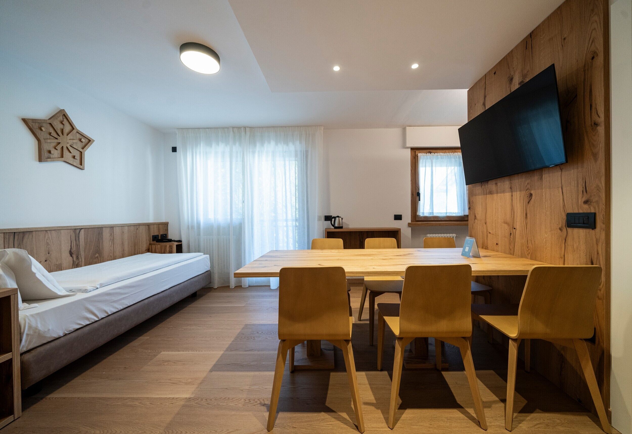 A modern room with a wooden floor, a sofa bed, a dining table with chairs, a flat-screen TV mounted on a wooden wall, and a window with curtains.