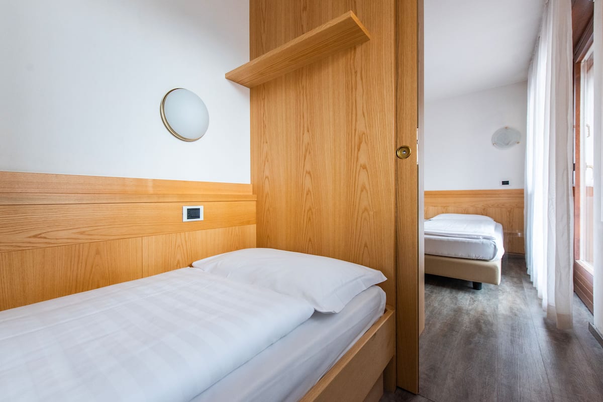 A single bed with a wooden headboard, a bedside table, a round mirror, and a small control panel on the wall.