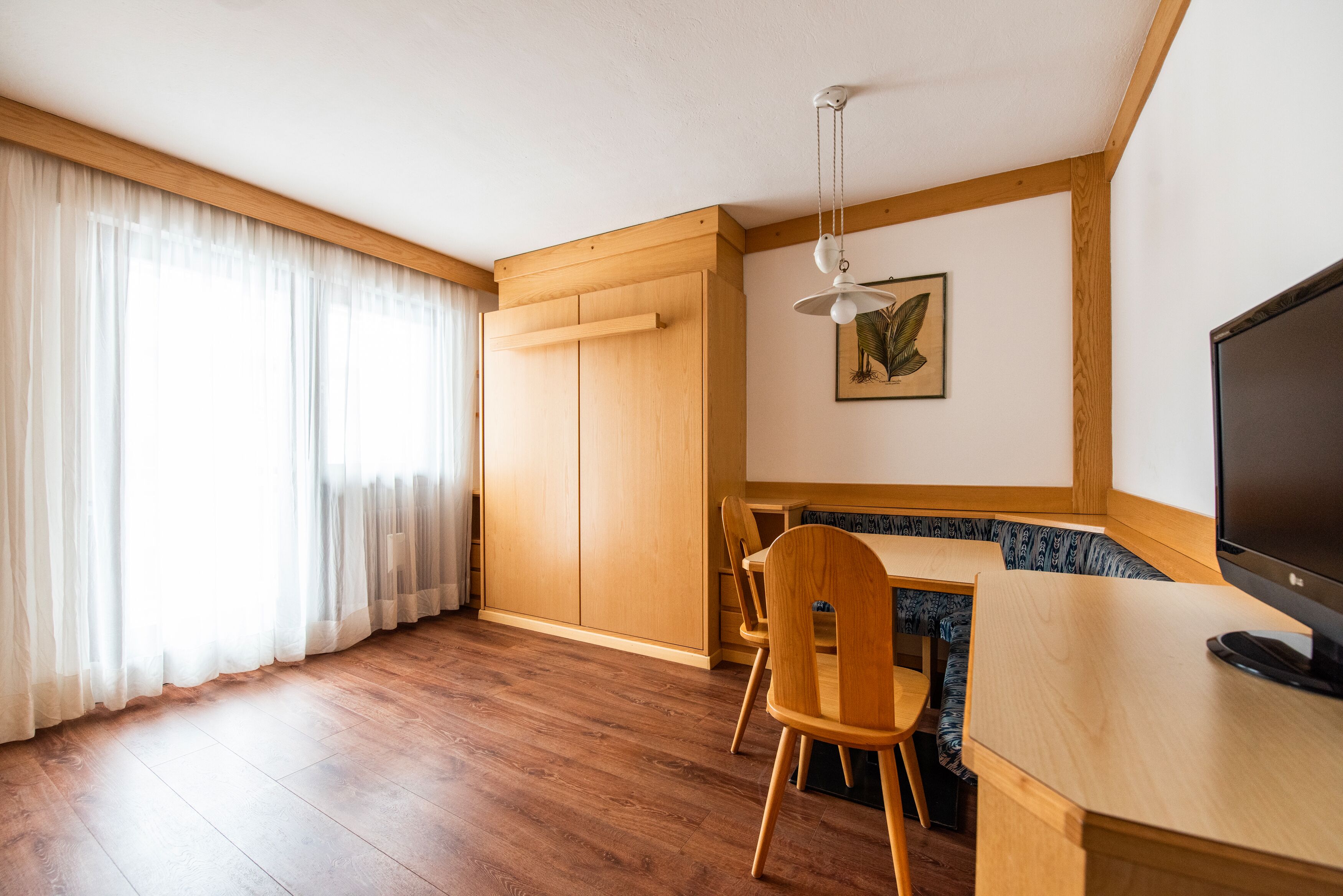 A room with wooden flooring, a dining table with chairs, a TV, and a window with curtains.