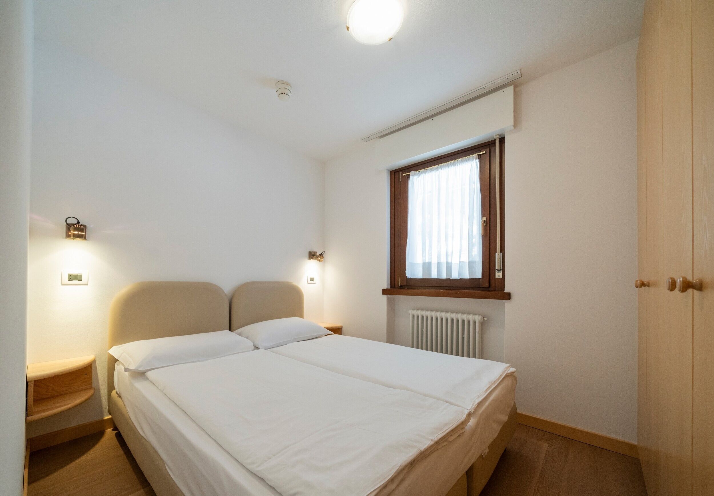 Une chambre à coucher avec un lit, des lampes de chevet, une fenêtre avec des rideaux, un radiateur et une armoire en bois.
