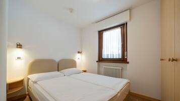 Une chambre à coucher avec un lit, des lampes de chevet, une fenêtre avec des rideaux, un radiateur et une armoire en bois.