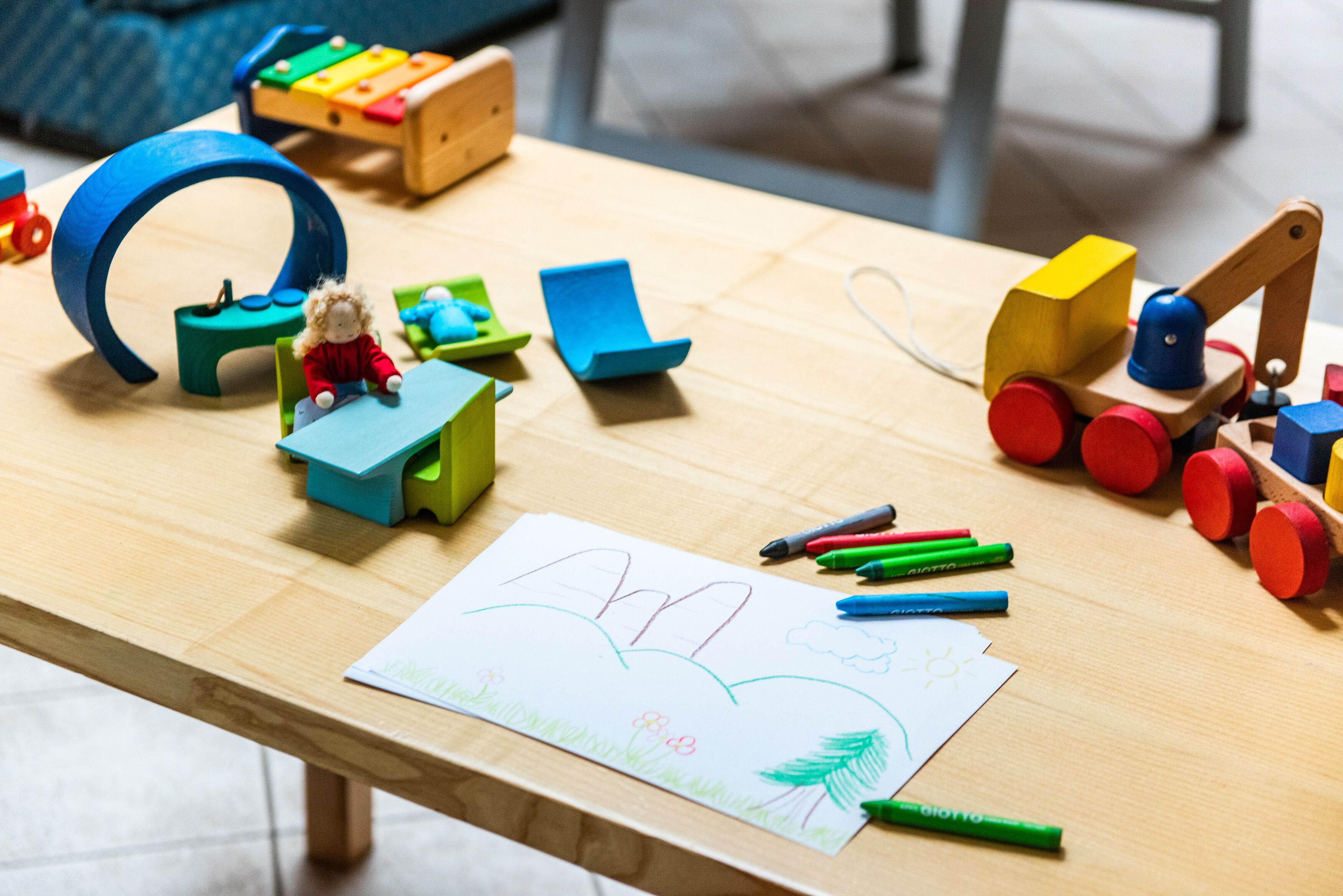 A wooden table with children's toys, a small doll, and a drawing paper with a drawing.
