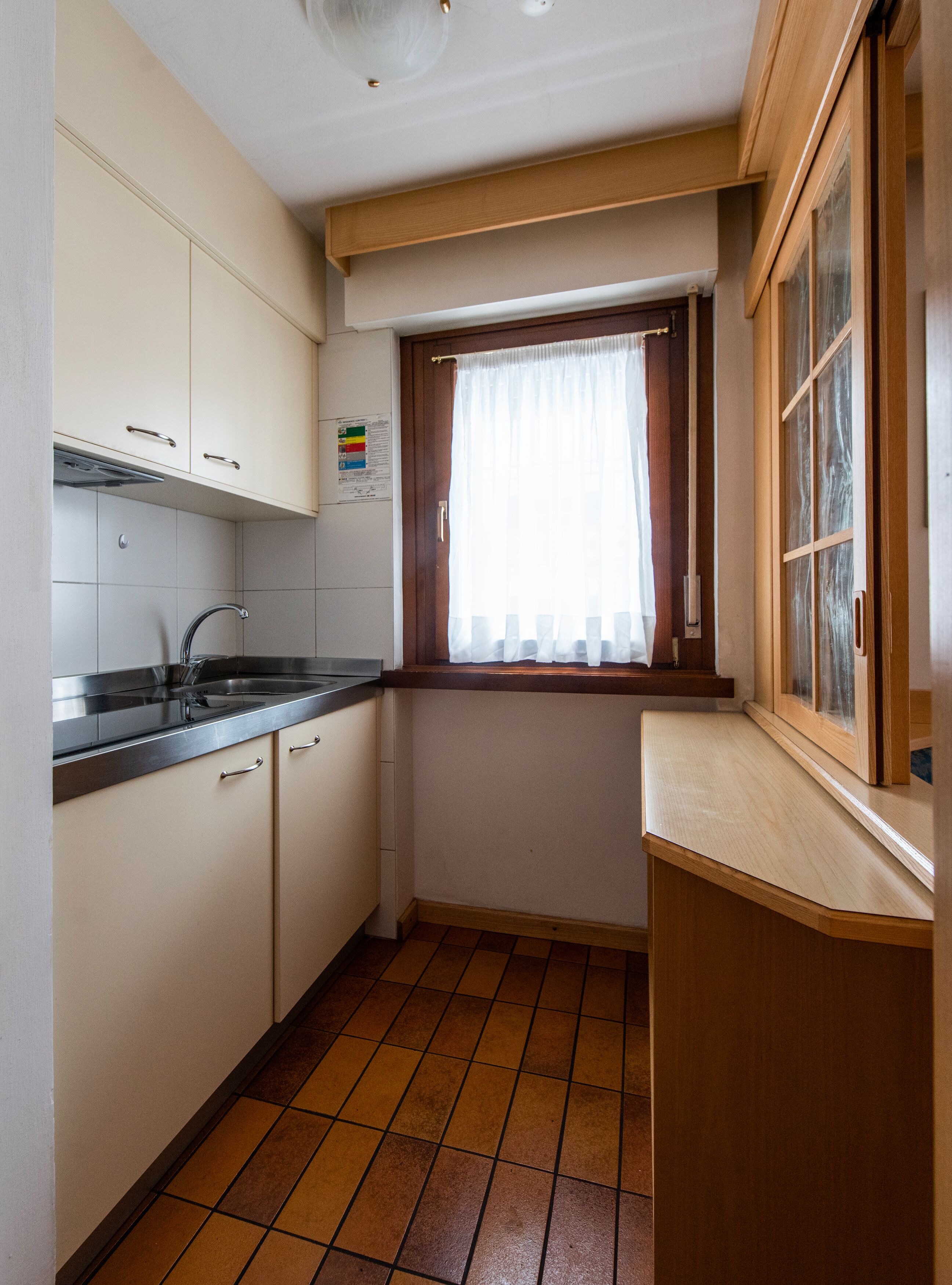 Cocina compacta con armarios blancos, un lavamanos de acero inoxidable y una ventana con marco de madera y cortinas blancas.