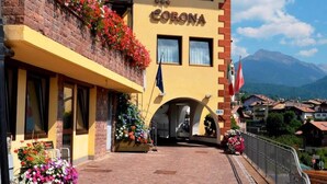 A hotel named "HOTEL CORONA" with a brick facade, flower boxes on the balcony, and a view of mountains in the background. - Corona (CARANO)