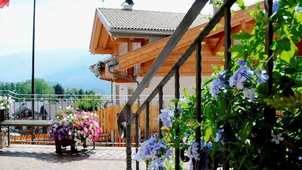 A wooden building with a sloped roof, a balcony, and a view of mountains in the background. - Corona (CARANO)