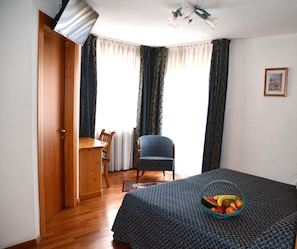 A room with a bed, a chair, a table, and a window with curtains. - Corona (CARANO)